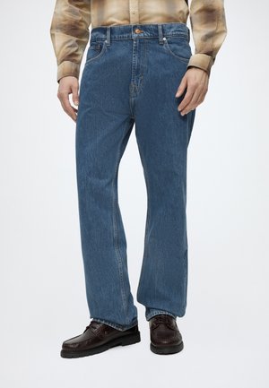 Mann trägt gerade geschnittene blaue Jeans, braune Lederschuhe und ein beigefarbenes Langarmhemd vor weißem Hintergrund.