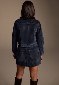 Veste en daim bleu marine avec jupe assortie, dotée de deux poches arrière et d'une coupe courte. Texture lisse et ajustée.
