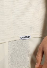 Beige idrottströja med ett strukturerat, andningsbart tyg och en Smilodox-logotyp vid fållen, med subtila mönsterdetaljer.