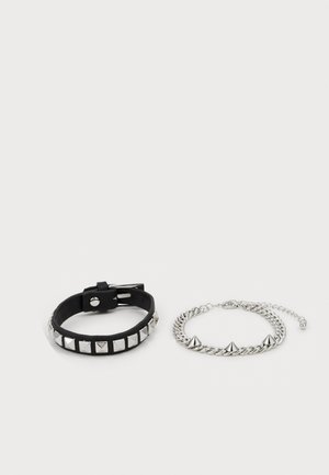 Pulseira de couro preto com pinos piramidais prateados e fivela ajustável, ao lado de uma pulseira de corrente prateada com três espigões salientes.