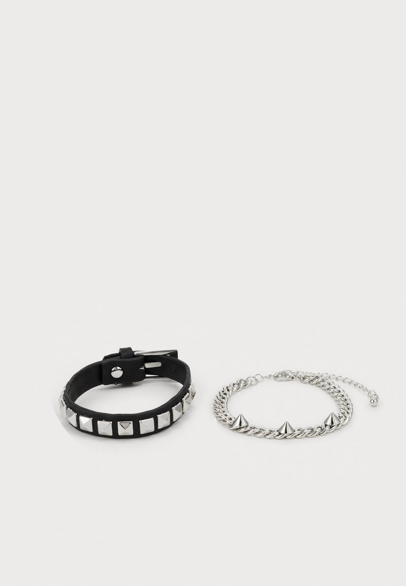 Bracelet en cuir noir avec des clous en pyramide argentés et une boucle ajustable, à côté d'un bracelet en chaîne argenté avec trois pointes saillantes.