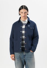 Navy blue jacket made of sturdy fabric, featuring a collared neckline, button front, two front pockets, and a chest pocket with a label.