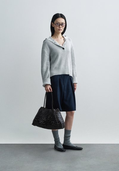 Grijze gebreide trui met een knoopsluiting bij de hals, marineblauwe knie-lange rok, getextureerde donkere tas, grijs gemêleerde sokken en zwarte platte schoenen.