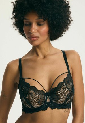 Žena s kučeravými vlasmi, ktorá má na sebe čiernu čipkovú podprsenku s kvetinovými vzormi a tenkými ramienkami, pozerá mierne nadol na jednofarebnom pozadí.