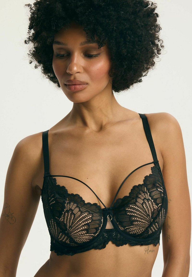 Žena s kučeravými vlasmi, ktorá má na sebe čiernu čipkovú podprsenku s kvetinovými vzormi a tenkými ramienkami, pozerá mierne nadol na jednofarebnom pozadí.
