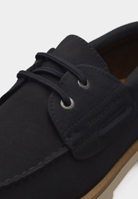 Μαύρο casual παπούτσι με κορδόνια, μεταλλικά οφθαλμικά, ορατή ραφή και γωνία σόλας ανοιχτό καφέ.