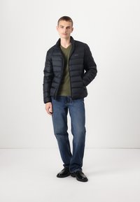 Giubbotto imbottito nero con colletto alto, chiusura con zip e cuciture orizzontali, abbinato a una t-shirt verde e jeans blu. Scarpe nere.