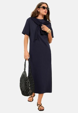 Frau trägt ein navyfarbenes Midikleid, passenden Pullover über die Schultern drapiert, schwarze Sonnenbrille, goldene Armbänder, schwarze Sandalen und hält eine schwarze geflochtene Tasche.