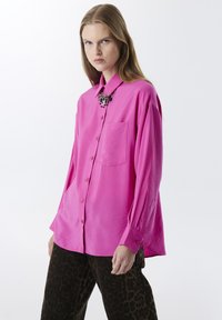Camisa de manga larga rosa brillante con diseño de botones y un bolsillo en el pecho. Combinada con pantalones de pierna ancha con estampado animal.
