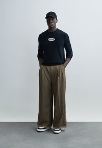 Μαύρο πουλόβερ με μακριά μανίκια και λογότυπο "VOLTAIRE", καφέ φαρδιά παντελόνια, μαύρο καπέλο και παπούτσια με λευκή σόλα. Μινιμαλιστικός σχεδιασμός, απαλό ύφασμα.