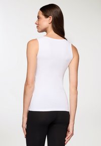 Donna con lunghi capelli castani, che indossa una maglietta bianca aderente senza maniche e pantaloni neri, di spalle contro uno sfondo chiaro e semplice.