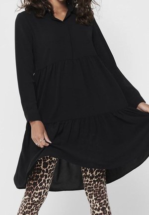 Woman wearing loose black long-sleeve dress lifting hem to show leopard print leggings beneath against plain background.