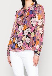 Blouse transparente à manches longues avec un imprimé floral multicolore et motif paisley sur fond noir, dotée d'un col froncé et de poignets boutonnés.