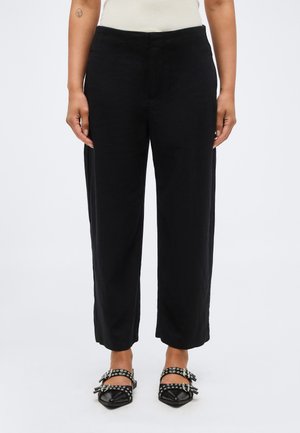 Femme portant un pantalon noir à jambes larges et des chaussures noires pointues avec des brides œillet, debout devant un fond blanc uni.