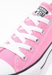 Růžové plátěné tenisky s bílými tkaničkami, stříbrnými očky a černobílou značkovou nálepkou na jazyku. Zakulacený gumičkový špička.