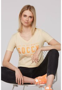 Camiseta de pico amarilla clara con "VIVE EN EL MOMENTO SOCXY REGISTRADO" en negrita naranja. El modelo lleva jeans negros y zapatillas naranjas.