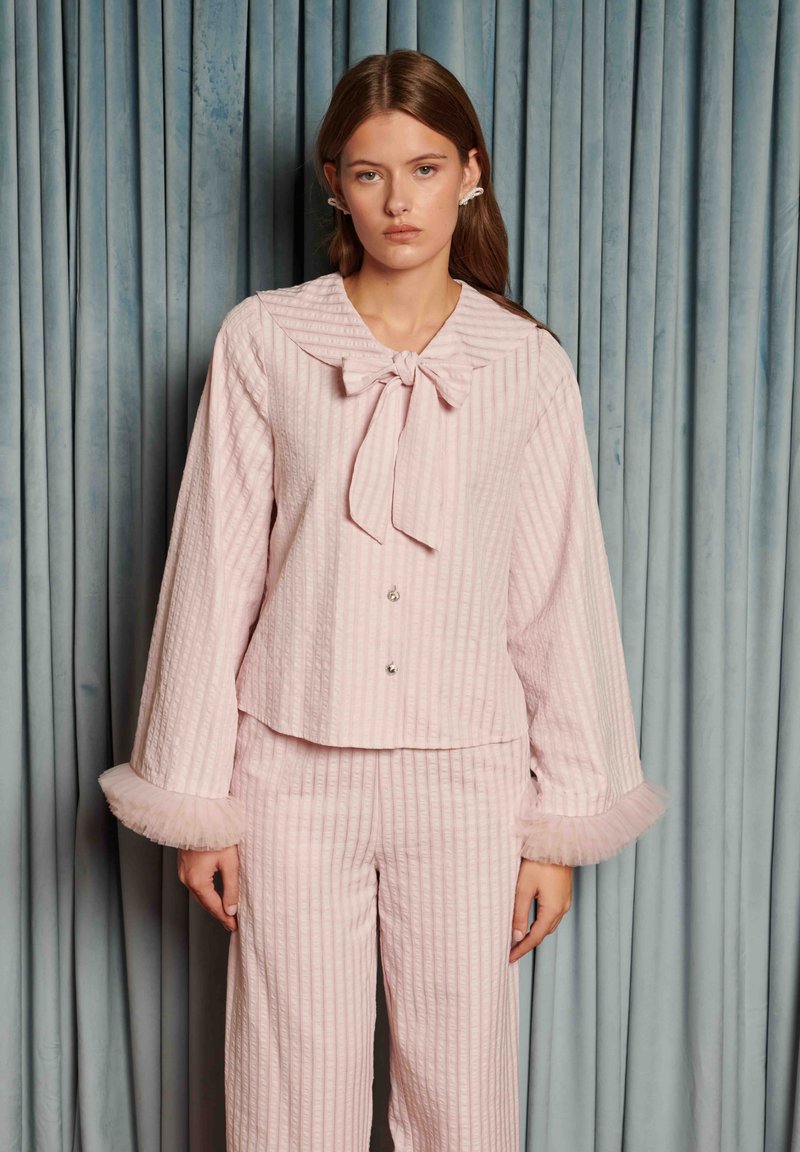 Ružový súbor na oddych pozostávajúci z košeľového vrchného dielu s golierom, zapínaného na gombíky a s mašľou, a širokých nohavíc, oba s textúrovanými pásikmi a tylovými ozdobami na manžetách.