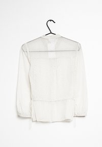 Blusa blanca translúcida con lunares negros, puños fruncidos, un lazo en la cintura y un cuello con solapa, colgando en un gancho negro.