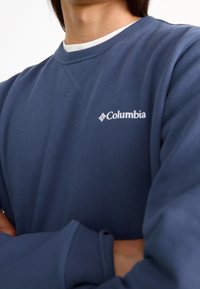 Granatowa bluza wykonana z mieszanki bawełny, z ribbikowymi mankietami i dekoltem, z białym logo Columbia na klatce piersiowej. Gładka tekstura.