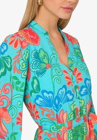 Abito camicia floreale in colori vivaci di turchese, rosso, rosa e verde. Presenta un colletto, bottoni e un dettaglio della vita legata.