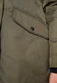Olivově zelená svrchní bunda s kapsou na klopu na knoflíky a strukturovanou tkaninou. Design zahrnuje minimalizované detaily prošívání a vroubkované manžety.
