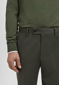 Man wearing dark green sweater tucked into dark gray tailored trousers with button and belt loops, hand relaxed by side.