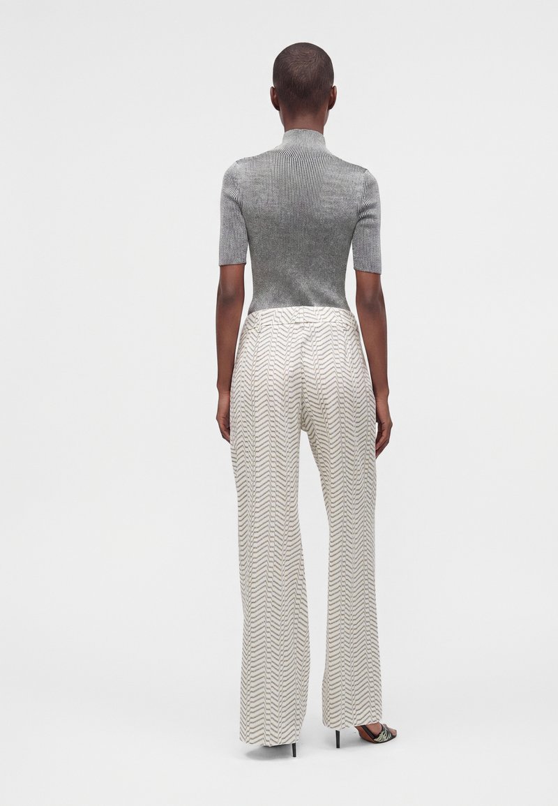 Personne portant un haut gris ajusté à manches courtes et un pantalon blanc taille haute à jambes larges avec un motif zigzag noir, debout de dos.