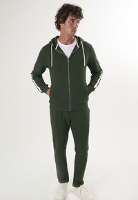 Groene zip-up hoodie met witte trekkoorden, gecombineerd met bijpassende groene joggers. Bevat witte zijstrepen en is gemaakt van zacht stof. Witte sneakers.