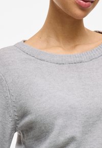 Gros plan d'une personne portant un pull léger gris clair en tricot avec un large col côtelé, montrant le cou et une partie du bas du visage.