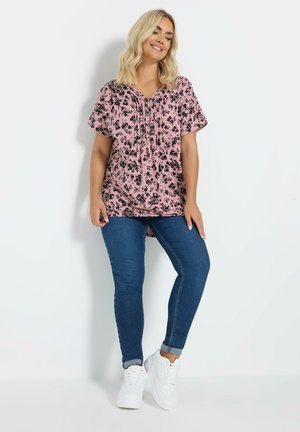 Roze top met korte mouwen, zwart bloemmotief, v-hals en geplooide details, gecombineerd met blauwe skinny jeans en witte sneakers.
