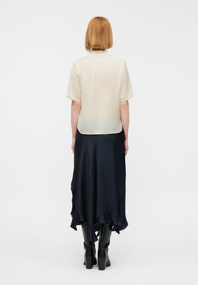 Κρεμ μεταξωτό πουκάμισο με κοντά μανίκια, συνδυασμένο με μακριά, ρέουσα φούστα σκούρου ναυτικού μπλε και μαύρες μπότες μέχρι το γόνατο. Λείες υφές, στρωμένο σχέδιο.