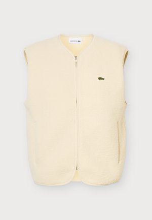 Un homme se tient de face, vêtu d’un gilet polaire crème zippé, d’un T-shirt vert, d’un pantalon beige et de baskets blanches, sur un fond uni.