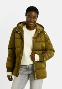 Doudoune verte olive avec capuche, dotée d'une fermeture éclair, de poignets côtelés et d'une texture brillante. Associée à un pull crème et à un jean gris.