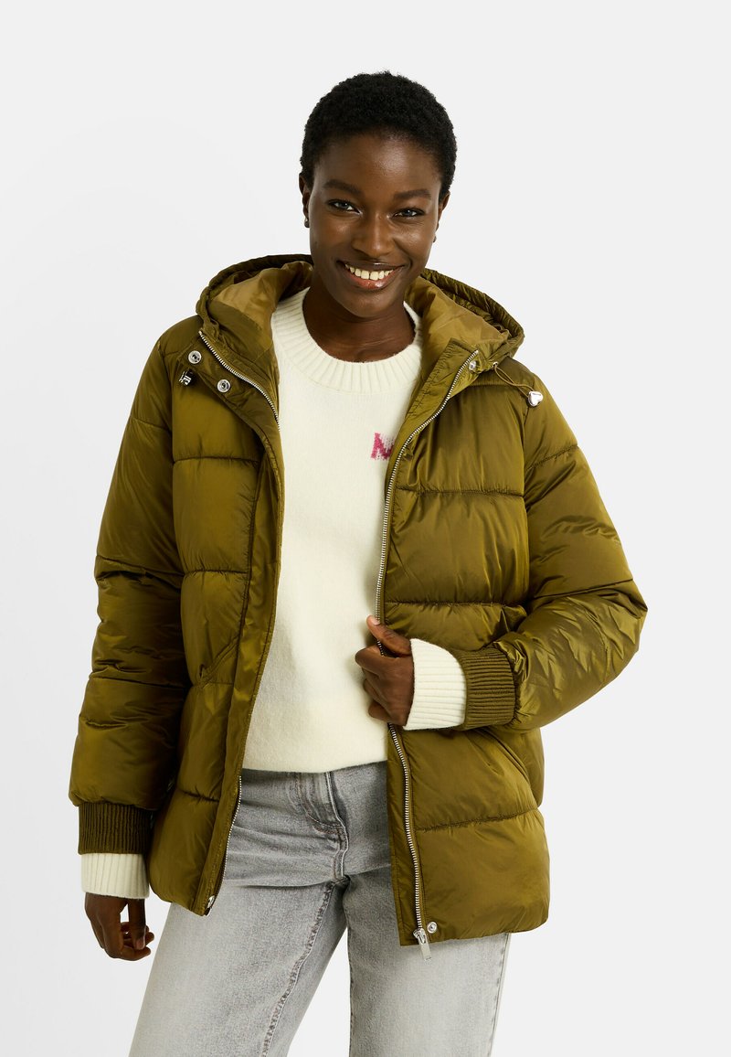 Doudoune verte olive avec capuche, dotée d'une fermeture éclair, de poignets côtelés et d'une texture brillante. Associée à un pull crème et à un jean gris.