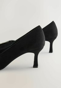 Chaussures à talons hauts en suédine noire avec un bout pointu et un talon élancé. Texture lisse avec un design de dos légèrement courbé.