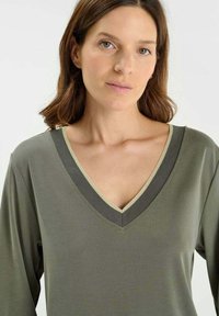 Woman with light skin and brown hair wearing an olive green V-neck top with dark and light trim, facing the camera against a plain background.