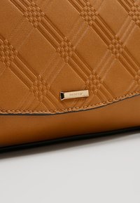 Gros plan d'une surface en cuir texturé fauve avec un motif à carreaux diagonaux et une petite étiquette métallique dorée rectangulaire.