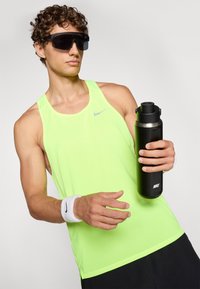 Jasnożółty sportowy podkoszulek z perforowaną teksturą, trzymający butelkę wody, czarne okulary przeciwsłoneczne, opaska na nadgarstek i ciemne szorty.