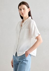 Camisa de linho branca de mangas curtas com colarinho notch, um bolso e um ajuste relaxado, combinada com jeans azul claro. Textura suave.