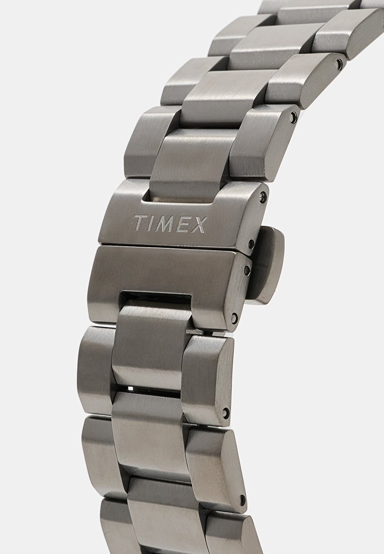 Correa de reloj plateada metálica con un cierre grabado con "TIMEX", que presenta eslabones de acero cepillado y un mecanismo de cierre plegable.