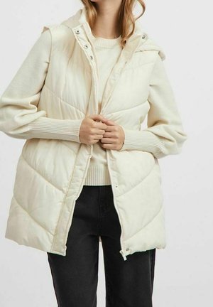 Osoba nosząca kremową pikowaną kamizelkę bez rękawów na dzianinowym swetrze, zapinającą zamek błyskawiczny, w połączeniu z czarnymi spodniami.