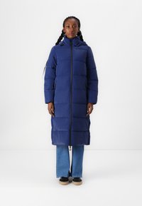 Geacă puffer lungă de culoare bleumarin, cu fermoar și glugă, având un design matlasat, buzunare laterale și o croială dreaptă, purtată cu blugi și adidași.