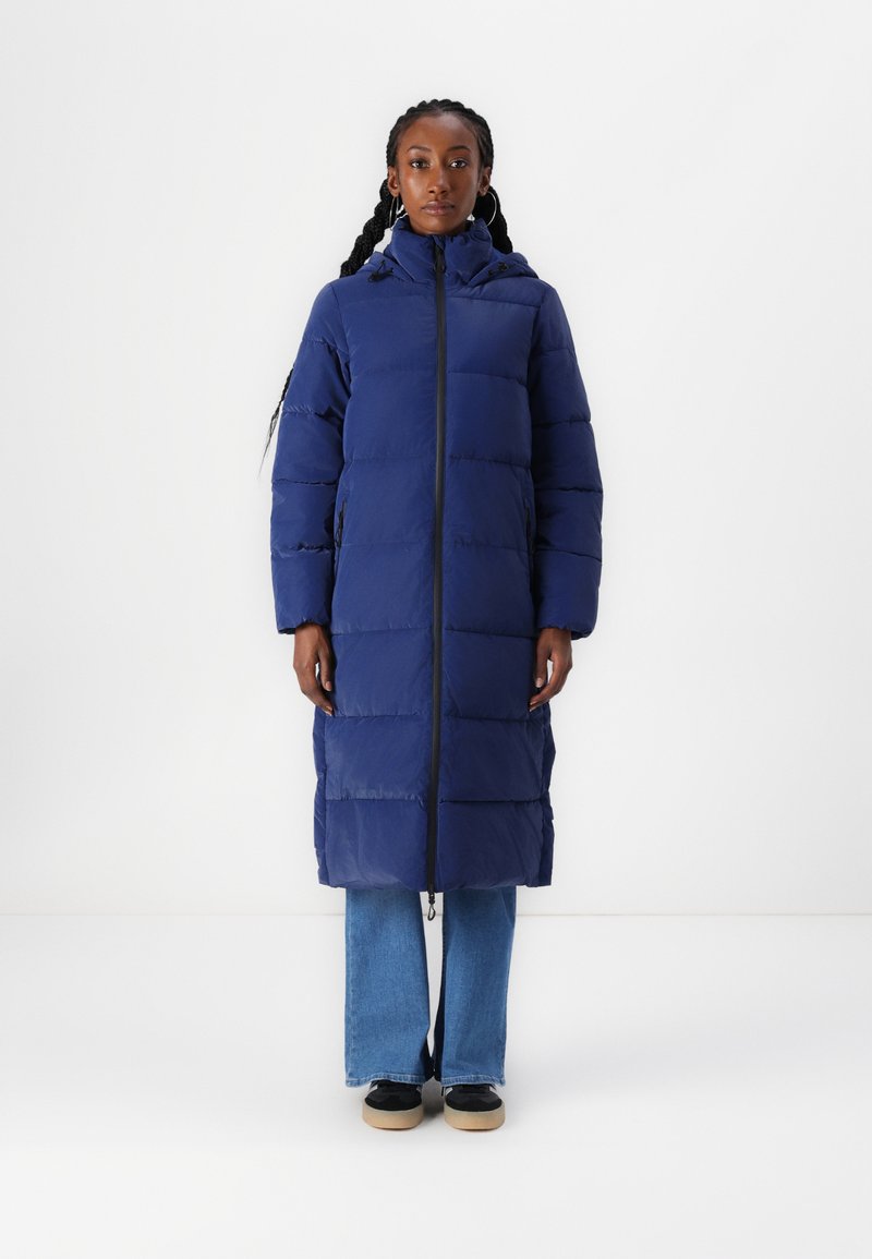 Geacă puffer lungă de culoare bleumarin, cu fermoar și glugă, având un design matlasat, buzunare laterale și o croială dreaptă, purtată cu blugi și adidași.