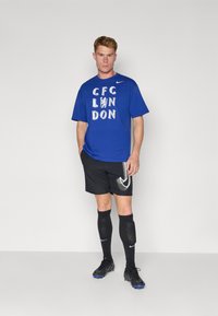 Blå kortærmet T-shirt med "CFC LONDON" i hvidt print, parret med sorte shorts med Nike-logo, sorte sokker og sorte fodboldsko.