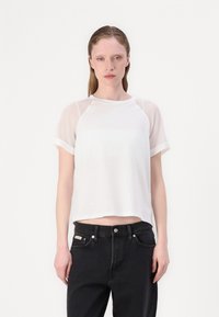 T-shirt bianco a maniche corte con la parte superiore in tessuto trasparente e la parte inferiore solida. Collo rotondo e vestibilità rilassata. Abbinato a jeans neri.