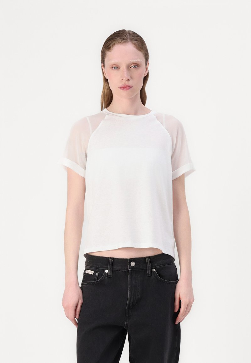 T-shirt bianco a maniche corte con la parte superiore in tessuto trasparente e la parte inferiore solida. Collo rotondo e vestibilità rilassata. Abbinato a jeans neri.