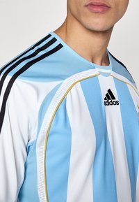 Αθλητική φανέλα Adidas με κατακόρυφες ρίγες σε ανοιχτό μπλε και λευκό, μαύρες λεπτομέρειες και στρογγυλή λαιμόκοψη με κίτρινα στοιχεία.