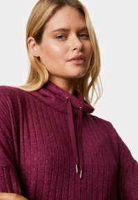 Purple ribbed pullover with a high neck and drawstring. Textured fabric with subtle shimmer. Features silver accents on drawstring ends.