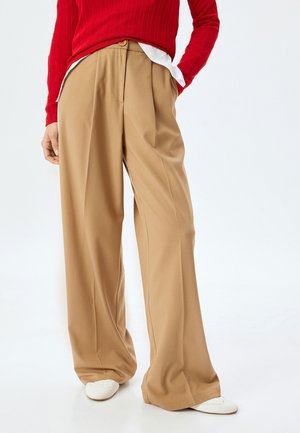 Beige wijde taillebroek met voorplooien en een enkele knoopsluiting, gecombineerd met een rode geribbelde trui. Horizontale textuur zichtbaar.