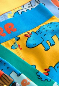 Gros plan sur des sous-vêtements d'enfants colorés avec des dinosaures bleus, des lettres orange, et des bandes de tissu jaune vif, bleu, vert et orange.